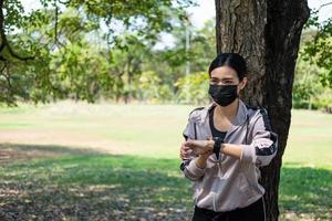 selettivo messa a fuoco a viso di giovane bellissimo asiatico donne indossare chirurgico viso maschera utilizzando inteligente orologio per traccia attività prima esercizio o in esecuzione a il parco nel il mattina. nuovo normale stile di vita. foto