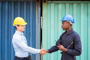 selettivo messa a fuoco a africano uomini lavoratore shake mano con caucasico nolo supervisore mentre Lavorando su luogo con sicurezza casco a contenitore logistica importare o esportare molo. industriale lavoro di squadra. foto