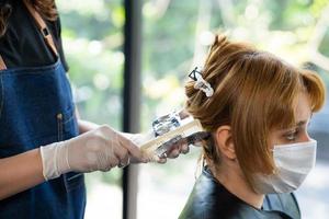 selettivo messa a fuoco a liquido capelli colore di capelli elegante mentre parrucchiere e mettere cura trattamento mentre messa in piega Riccio capelli per cliente. professionale occupazione, bellezza e moda servizio durante covid-19 foto