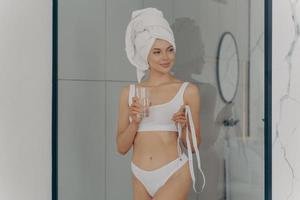 adatta bella ragazza in bagno con un bicchiere di acqua fresca e nastro di misurazione foto