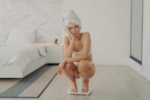 la giovane ragazza europea triste frustrata si accovaccia sulla bilancia in camera da letto dopo la doccia quotidiana foto