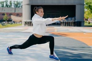 foto all'aperto di snella donna sportiva coinvolta in attività ricreative allunga braccia gambe guarda in lontananza indossa leggings bianchi con cappuccio e scarpe sportive stende il corpo prima di correre conduce uno stile di vita sano