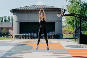 giovane donna atletica in abbigliamento sportivo nero salta in alto tiene le braccia alzate fibbie le mani si riscalda prima di fare jogging si allena allo stadio all'aperto ha cardio brucia grassi e calorie per dimagrire foto