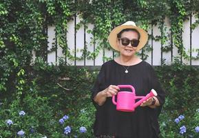 contento e salutare anziano asiatico donna indossare nero camicetta, occhiali da sole e cappello, Tenere rosa irrigazione può nel il giardino, sorridente e guardare a telecamera. foto