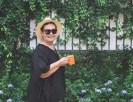 salutare anziano asiatico donna indossare nero camicetta, occhiali da sole e cappello, Tenere arancia colore tazza di caffè nel il giardino con viola fiori, sorridente e guardare a telecamera. foto