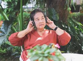contento e salutare asiatico anziano donna seduta con caffè tazza e fiore pentole nel all'aperto giardino, ascoltando per preferito musica a partire dal cuffia, sorridente e guardare lontano. foto