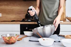 donna nel cucina cucinando un' torta. mani battere il Impasto con un elettrico miscelatore foto