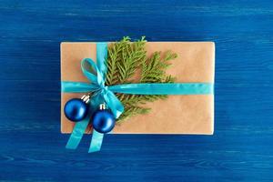 regalo scatola avvolto di mestiere carta, blu nastro e decorato abete rami e blu Natale palle su il blu di legno sfondo, superiore Visualizza. Natale regalo. foto