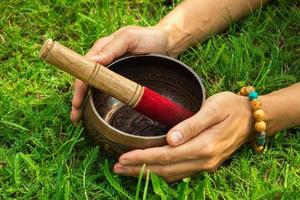 il mani di un' giovane donna su tibetano cantando ciotola con di legno bastone su il erba. foto