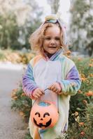 carino poco ragazza nel un' arcobaleno unicorno costume per Halloween va per raccogliere dolci nel un' zucca cestino nel un' Residenziale la zona. bambino passeggiate nel il all'aperto. trucco o trattare. stile di vita. kigurumi foto