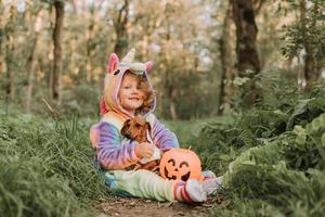 poco ragazza nel un' arcobaleno unicorno Halloween costume e un' bassotto nel un' vestito con un' zucca cestino per dolci siamo seduta su ceppo a foresta tramonto. favoloso meraviglioso magico foresta. spazio per testo foto