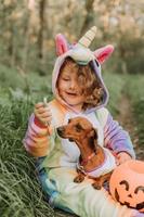 poco ragazza nel un' arcobaleno unicorno Halloween costume e un' bassotto nel un' vestito con un' zucca cestino per dolci siamo seduta su ceppo a foresta tramonto. favoloso meraviglioso magico foresta. spazio per testo foto