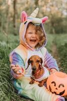 poco ragazza nel un' arcobaleno unicorno Halloween costume e un' bassotto nel un' vestito con un' zucca cestino per dolci siamo seduta su ceppo a foresta tramonto. favoloso meraviglioso magico foresta. spazio per testo foto