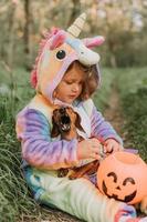 poco ragazza nel un' arcobaleno unicorno Halloween costume e un' bassotto nel un' vestito con un' zucca cestino per dolci siamo seduta su ceppo a foresta tramonto. favoloso meraviglioso magico foresta. spazio per testo foto