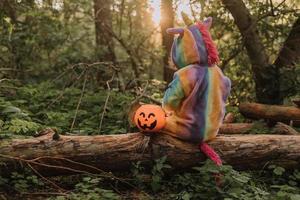 poco ragazza nel un' arcobaleno unicorno Halloween costume con un' zucca cestino per dolci è seduta su un' ceppo a un' foresta tramonto. un' favoloso meraviglioso magico foresta. spazio per testo. alto qualità foto