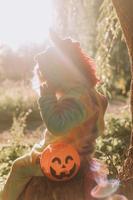 poco ragazza nel un' arcobaleno unicorno Halloween costume e un' bassotto nel un' vestito con un' zucca cestino per dolci siamo seduta su ceppo a foresta tramonto. favoloso meraviglioso magico foresta. spazio per testo foto