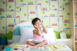 asiatico bambini utilizzando cuffie per ascolta musica di smartphone su il letto nel sua decorato Camera da letto foto