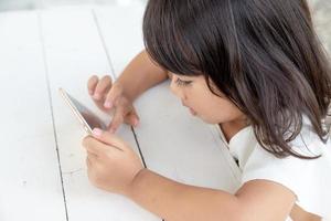 asiatico ragazza giocando smartphone su il tavolo. Guardando smartphone. bambini uso telefoni e giocare Giochi. bambino uso mobile. avvincente gioco e cartone animato foto