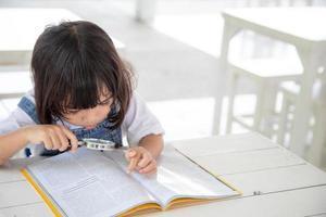 asiatico poco ragazza lettura il libri su il scrivania con un' ingrandimento bicchiere foto