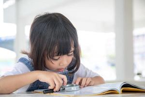 asiatico poco ragazza lettura il libri su il scrivania con un' ingrandimento bicchiere foto