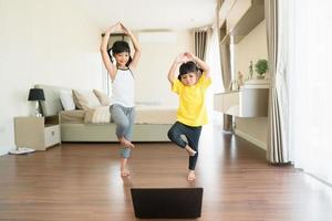 Due poco ragazze praticante yoga, allungamento, fitness di video su taccuino. lontano in linea formazione scolastica addestramento, aerobico a casa. salutare stile di vita, coronavirus, restare casa. bambini sport casa quarantena. foto