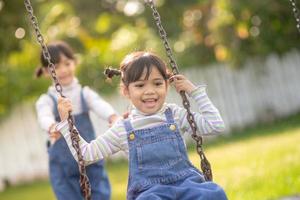 Due carino poco sorelle avendo divertimento su un' swing insieme nel un' bellissimo estate giardino su un' caldo e soleggiato giorno all'aperto. attivo estate tempo libero per bambini. foto