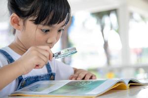 asiatico poco ragazza lettura il libri su il scrivania con un' ingrandimento bicchiere foto