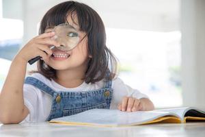 asiatico poco ragazza lettura il libri su il scrivania con un' ingrandimento bicchiere foto