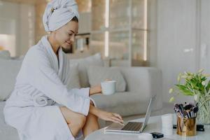 ripresa in interni di freelancer femminile lavora lontano da casa tipi sul computer portatile vestito in accappatoio beve caffè o tè gode dell'atmosfera domestica naviga in Internet legge consigli sulla cura della pelle foto