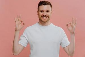 il giovane uomo barbuto soddisfatto fa un gesto ok con entrambe le mani esprime accordo approva qualcosa elogia un bel lavoro dice ben fatto vestito con una maglietta bianca casual isolata sulla parete rosa dello studio foto