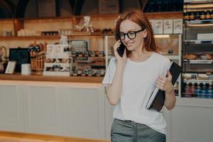 giovane donna rossa piacevole freelance che parla al telefono e sorride mentre lascia la caffetteria foto