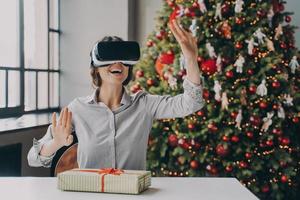 sorpresa giovane donna che indossa un gadget auricolare vr seduto al tavolo con confezione regalo di natale avvolta foto