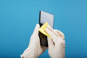 mani nel protettivo guanti pulito e disinfettare un' smartphone con un' giallo tovagliolo su un' blu sfondo. foto