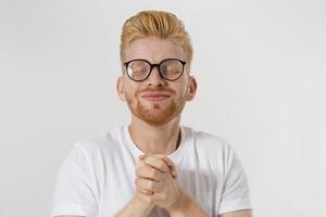 pregando giovane isolato su sfondo bianco. copia spazio. esprimere un desiderio e un concetto di fortuna. ragazzo dai capelli rossi con barba rossa e occhiali in maglietta bianca vuole che i sogni diventino realtà. successo e buona fortuna. foto