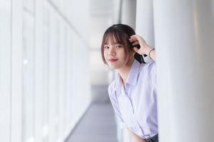 bellissimo alto scuola asiatico alunno giovane ragazza nel il scuola uniforme sta e sorrisi felicemente mentre lei con fiducia sembra a il telecamera con il scuola edificio come un' sfondo. foto