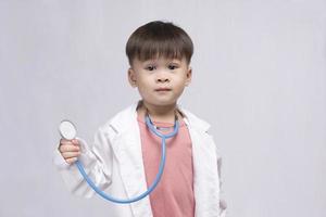 giovane carino asiatico ragazzo indossare un' medico uniforme Tenere un' stetoscopio giocando contento medico su bianca sfondo. prescolastico bambini fare finta per essere un' pediatra. foto