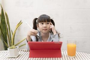 asiatico ragazza studiando in linea su sua scrivania a casa con tavoletta e penna. homeschooling durante quarantena. restare a casa intrattenimento. bambini e tavoletta, bambini siamo pensiero e analizzando compiti a casa. foto