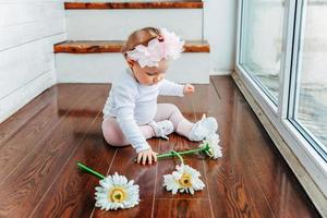 poco sorridente bambino ragazza uno anno vecchio indossare primavera ghirlanda ubicazione su pavimento nel luminosa leggero vivente camera vicino finestra e giocando con gerbera fiori. contento ragazzo giocando a casa. infanzia concetto. foto