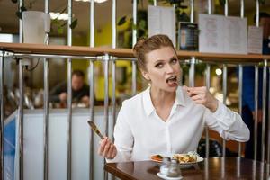 un' donna nel un' bianca camicia è seduta a un' tavolo mangiare con un' coltello e forchetta nel sua mani foto