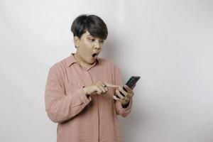 sorpreso capelli corti asiatico donna indossare un' rosa camicia puntamento a sua smartphone, isolato di un' bianca sfondo foto