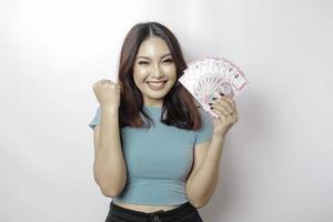 un' contento giovane donna è indossare blu maglietta e Tenere denaro contante i soldi nel indonesiano rupia isolato di bianca sfondo foto