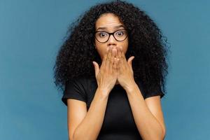 la foto di una giovane donna stupefatta copre la bocca, ha il respiro affannoso, gli occhi stropicciati, i capelli afro, non riesce a credere a notizie scioccanti, indossa una maglietta nera casual, si trova su sfondo blu. omg concetto.