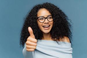 ritratto di donna felice con acconciatura afro, mostra il pollice in su, dà un'opinione positiva, guarda da parte, indossa occhiali ottici e maglione, isolato su sfondo blu. persone, accordo, approvazione foto