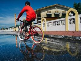 un' ragazzo nel rosso equitazione il bicicletta in il acqua pozzanghera su il suo modo per il moschea foto