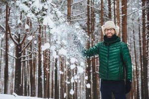 sorridente delizioso maschio vestito con abiti caldi, si trova nella foresta invernale, lancia la neve in aria, si diverte da solo, ha buon umore, esprime emozioni e sentimenti positivi. concetto di positività foto
