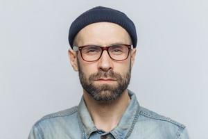 foto di un uomo elegante e sicuro di sé intelligente con barba e baffi scuri e folti, guarda seriamente nella fotocamera, posa su sfondo grigio dello studio, indossa occhiali e cappello. concetto di espressioni facciali