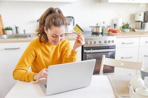 donna shopping in linea e pagare con oro credito carta. giovane ragazza seduta con il computer portatile acquisto su Internet accedere credito carta dettagli su cucina interno sfondo. in linea shopping e-commerce concetto. foto