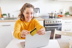 donna shopping in linea e pagare con oro credito carta. giovane ragazza seduta con il computer portatile acquisto su Internet accedere credito carta dettagli su cucina interno sfondo. in linea shopping e-commerce concetto. foto