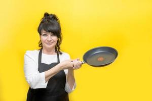 sorridente bella anziano donna cucinare nel nero cucina grembiule Tenere un' frittura padella nel sua mani, giallo sfondo foto