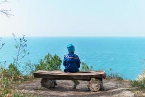 un' solitario bambino è riposo su un' panchina nel il foresta con un' Visualizza di il mare. foto